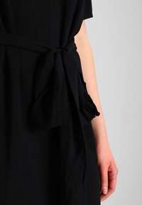 Närbild av en persons hand bredvid ett knutet svart tygbälte på ett svart plagg mot en enkel ljus bakgrund.