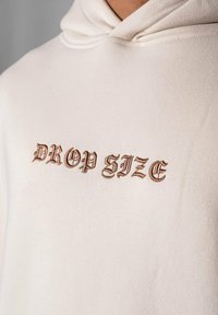 Beige Kapuzenpullover aus weichem Material mit einer vorderen Känguru-Tasche. Ausgestattet mit der aufgestickten Aufschrift "DROP SIZE" in brauner Schrift. Mit Kapuzendesign.