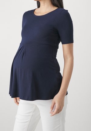 Pregnant woman wearing a fitted navy blue short-sleeve top and white pants, standing against a plain light background.