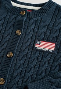 Jūras spēku krāsas adīts kardigans ar ribotu apkakli, brūniem pogām un uzlīmi ar ASV karogu un "SUPERDRY" logotipu.