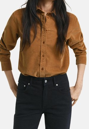 Persona vistiendo una camisa marrón de pana de manga larga con botones y vaqueros negros de cintura alta, con las manos descansando cerca de los bolsillos.