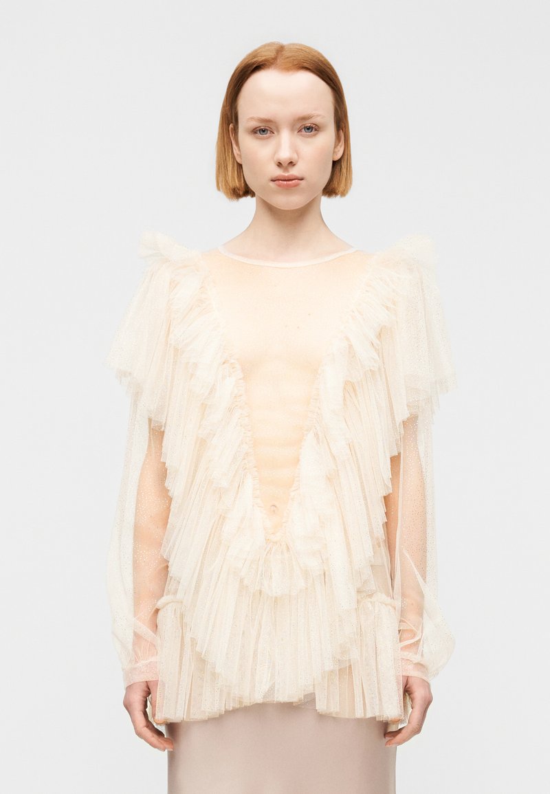 Νεαρό άτομο με ανοιχτόχρωμο δέρμα και κοντά κόκκινα μαλλιά, φοράει διάφανο μπεζ τοπ με στρωματοποιημένα βολάν και μακριά μανίκια, κοιτάζοντας μπροστά.