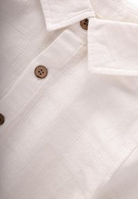 Camicia bianca con bottoni frontali, caratterizzata da un motivo a quadri testurizzato, bottoni in legno e un design del colletto classico. Il tessuto sembra leggero e tessuto.