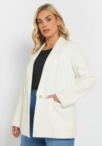 Weißer strukturierter Blazer mit rever collar, einem Knopfverschluss und Fronttaschen, kombiniert mit einem schwarzen Oberteil und blauen Jeans.