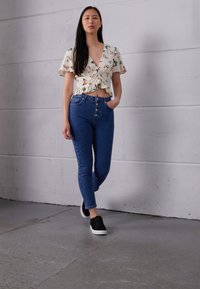 Top branco floral com bainha franzida, combinado com jeans azuis de cintura alta com cinco botões. Ténis pretos completam o visual.