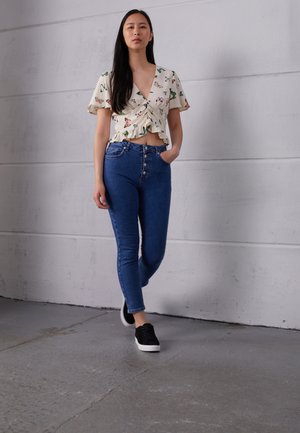 Top branco floral com bainha franzida, combinado com jeans azuis de cintura alta com cinco botões. Ténis pretos completam o visual.
