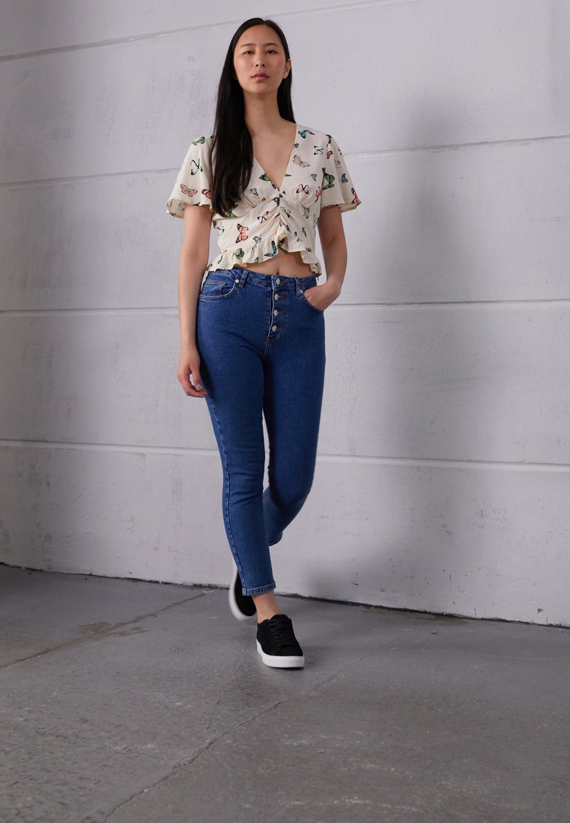 Top branco floral com bainha franzida, combinado com jeans azuis de cintura alta com cinco botões. Ténis pretos completam o visual.
