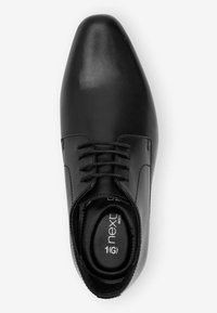 Chaussure de ville en cuir noir avec un bout pointu, fermeture à lacets, intérieur rembourré et coupe large. Texture lisse avec des accents minimaux.