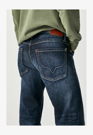 Tumedast sinisest denimist sirge lõikega teksapüksid, millel on kaks tagumist taskut koos õmblustega ja nahast logo silt vöökoha juures.
