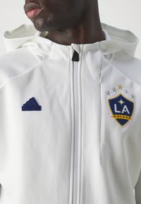 Witte zip-up jas met een capuchon, voorzien van een blauw en geel LA Galaxy-logo, een zwart Adidas-embleem en een gladde textuur.