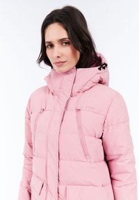 Mujer vistiendo una chaqueta de invierno acolchada de color rosa claro con un cuello alto, capucha y bolsillos con cremallera en el pecho, frente a un fondo blanco.