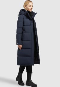 Lang navy pufferjas met capuchon, gequilt ontwerp, ritssluiting aan de voorkant en zijzakken. Gecombineerd met zwarte enkelboots.