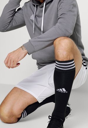 Felpa grigia con cappuccio e lacci bianchi, logo bianco, pantaloni corti bianchi e calze nere con strisce bianche. Scarpe da calcio nere visibili in basso.