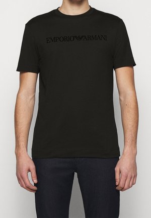 Czarna koszulka z krótkim rękawem z logo "Emporio Armani" w ciemnym nadruku na klatce piersiowej, noszona przez osobę z widocznymi ramionami i dłońmi.