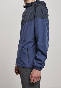 Marinblå vindjacka med en kontrasterande mörkblå övre panel, justerbar huva, ficka med dragkedja på sidan och elastiska ärmslut. Slätt, lätt material.