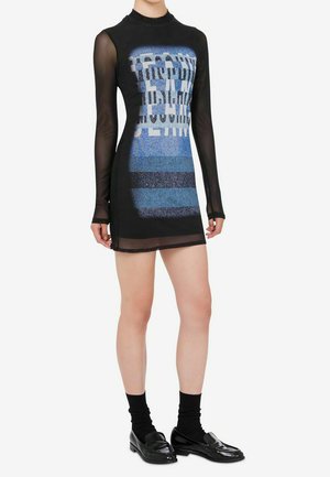 Mini abito nero in mesh aderente a maniche lunghe con scritta grafica blu sul davanti, indossato con calzini neri e mocassini neri lucidi.