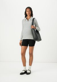 Svart och vit randig långärmad polo skjorta, svarta shorts, svart handväska, vita strumpor, svarta loafers med guldackcenter, stående pose.