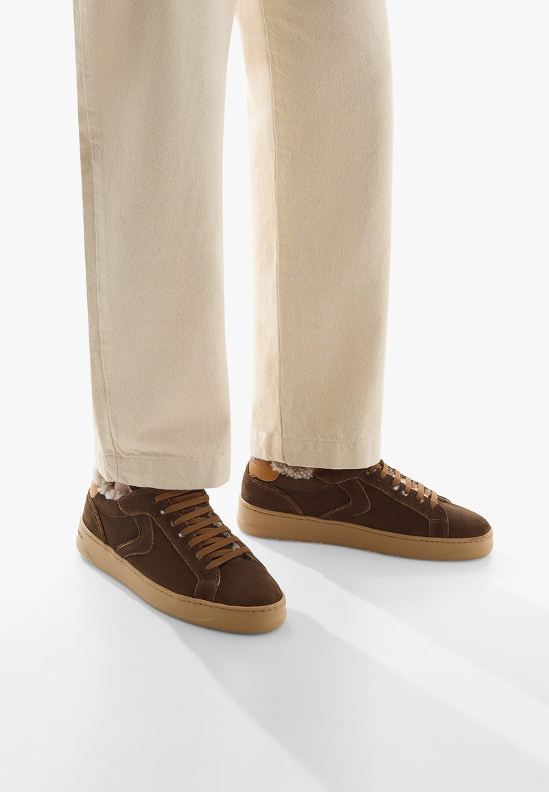 Sneaker in suede marrone con suole e lacci in gomma beige. Accenti in pelle tan evidenti e foderatura morbida visibile. I pantaloni sono beige, leggermente accorciati.