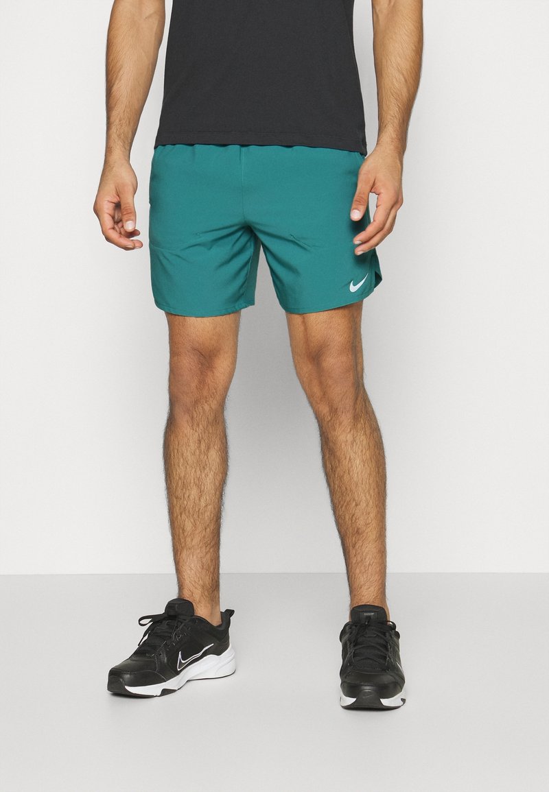Man iklädd turkosa Nike träningsshorts, svarta Nike-sneakers och en svart T-shirt, som står mot en ren vit bakgrund.