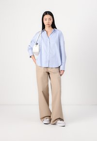 Light blue and white striped button-up shirt, beige wide-leg pants, and white sneakers; model holds a small white bag.