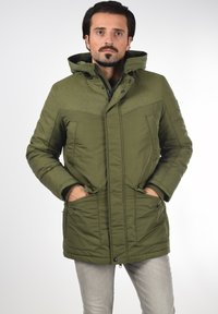 Parka verde con una sección superior texturizada, dos bolsillos frontales y una capucha. Cuenta con cierre de cremallera y detalles de costura visibles.