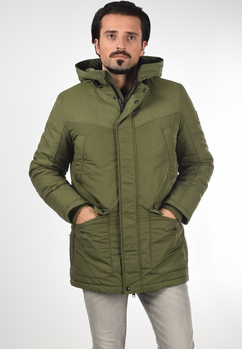 Parka verde con una sección superior texturizada, dos bolsillos frontales y una capucha. Cuenta con cierre de cremallera y detalles de costura visibles.