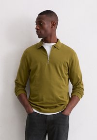 Olivgrüner, langärmeliger, mit Reißverschluss versehenen Pullover. Glattes Material mit einem kragenlosen Halsauschnitt, über einem weißen T-Shirt getragen, kombiniert mit dunklen Hosen.