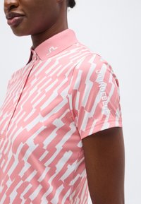 Γυναίκα που φοράει ροζ και λευκό κοντομάνικο πόλο με μοτίβο και κεντημένο λογότυπο στο γιακά και το μανίκι.