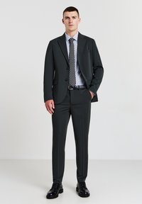 Dunkelgrauer Anzug mit einer geknöpften Jacke, kombiniert mit einem hellblauen gestreiften Hemd und einer gemusterten Krawatte. Schwarze Schuhe runden das Outfit ab.