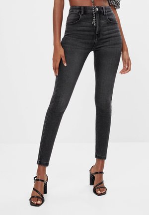 Kvinde iført højtaljede sorte skinny jeans og sorte sandaler med blokhæl og stropper, stående med en hånd hvilende på låret.