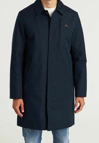 Un hombre lleva un abrigo largo azul marino con bolsillos en la parte delantera, un pequeño parche en el pecho y jeans azules, de pie contra un fondo claro y liso.