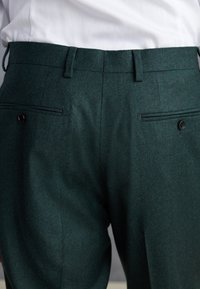 Pantaloni verdi scuro su misura con tasche posteriori a filetto fissate da bottoni neri, indossati con una camicia bianca infilata dentro.