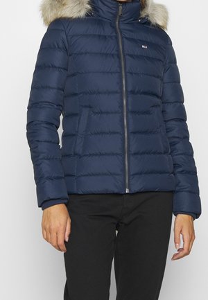 Veste matelassée bleu marine avec fermeture éclair à l'avant et capuche bordée de fourrure, portée avec un pantalon noir, vue du cou aux hanches.