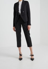 Donna che indossa pantaloni neri sartoriali corti, blazer nero doppiopetto, camicia bianca e décolleté nere con dettaglio a fiocco.