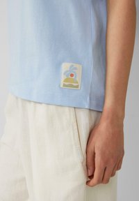 Lichtblauwe t-shirt met korte mouwen en een gestikt vierkant patch met een kleurrijk ontwerp van een palm en zon aan de linksonderzijde.