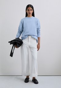 Sudadera azul claro con puños acanalados, pantalones blancos de pierna ancha con dobladillos enrollados y bolso de cuero negro. Zapatos de tela con textura completan el look.