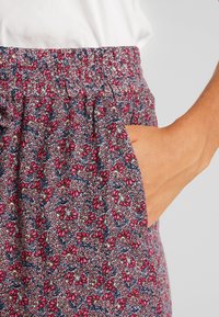 Pantalon à imprimé floral avec taille élastique, présentant une base bleu marine avec des fleurs rouges et vertes. Comprend un détail de poche latérale.