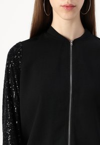 Schwarze Zip-Jacke mit glattem Stoffkörper und mit Pailletten verzierten Ärmeln. Verfügt über einen runden Halsausschnitt und silberne Reißverschlussdetails.