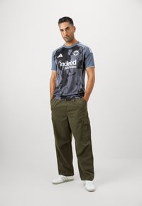 Camiseta de fútbol de patrón camuflado en negro y gris con mangas cortas, que presenta rayas blancas y el logo "indeed". Combinada con pantalones cargo de color verde oliva.