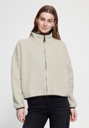 Jeune femme aux longs cheveux bruns portant une veste polaire beige clair zippée et un pantalon noir sur un fond clair.