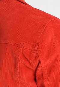 Giacca di tessuto corduroy rosso con superficie testurizzata, dotata di cucitura sulle spalle e di un colletto. Nessun bottone o tasca visibile.