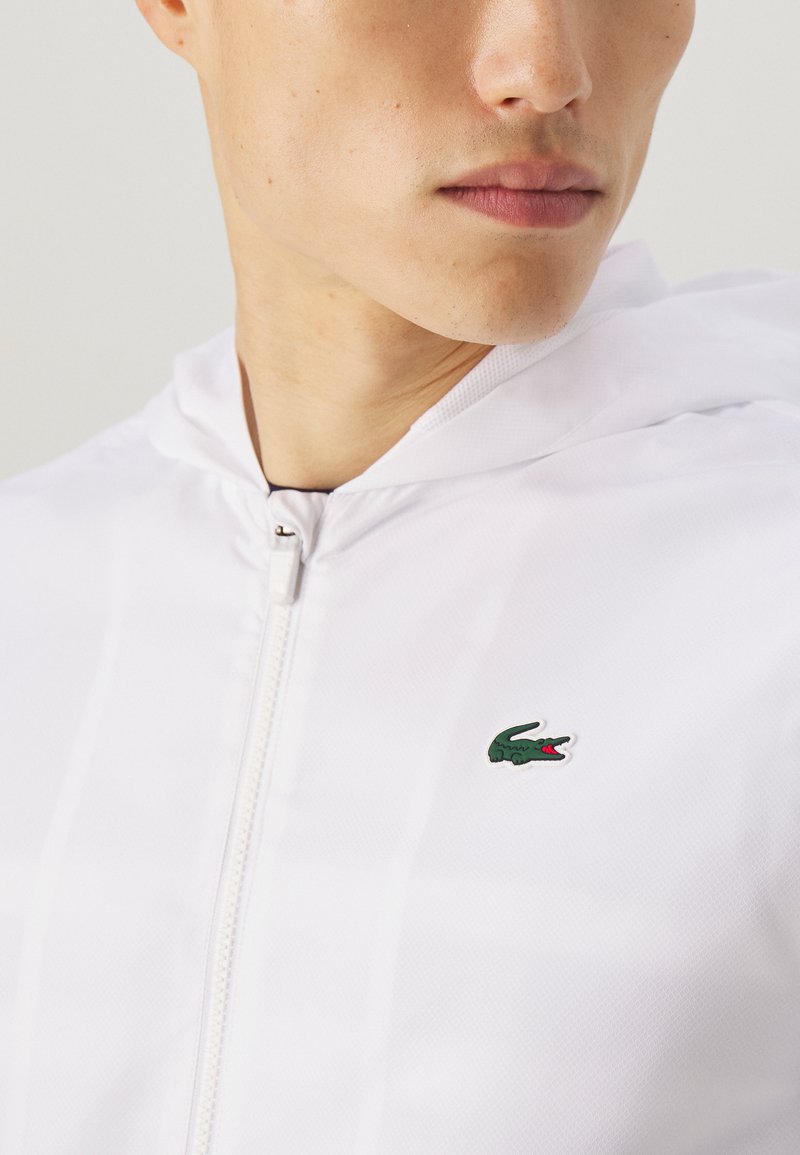 Chaqueta blanca con cremallera hecha de una tela ligera, que cuenta con un pequeño logo de cocodrilo verde en el lado izquierdo del pecho, con capucha y textura suave.