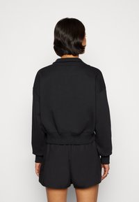 Vrouw met kort donker haar, gekleed in een zwart sweatshirt met lange mouwen en zwarte korte broek, gezien van achteren tegen een effen achtergrond.