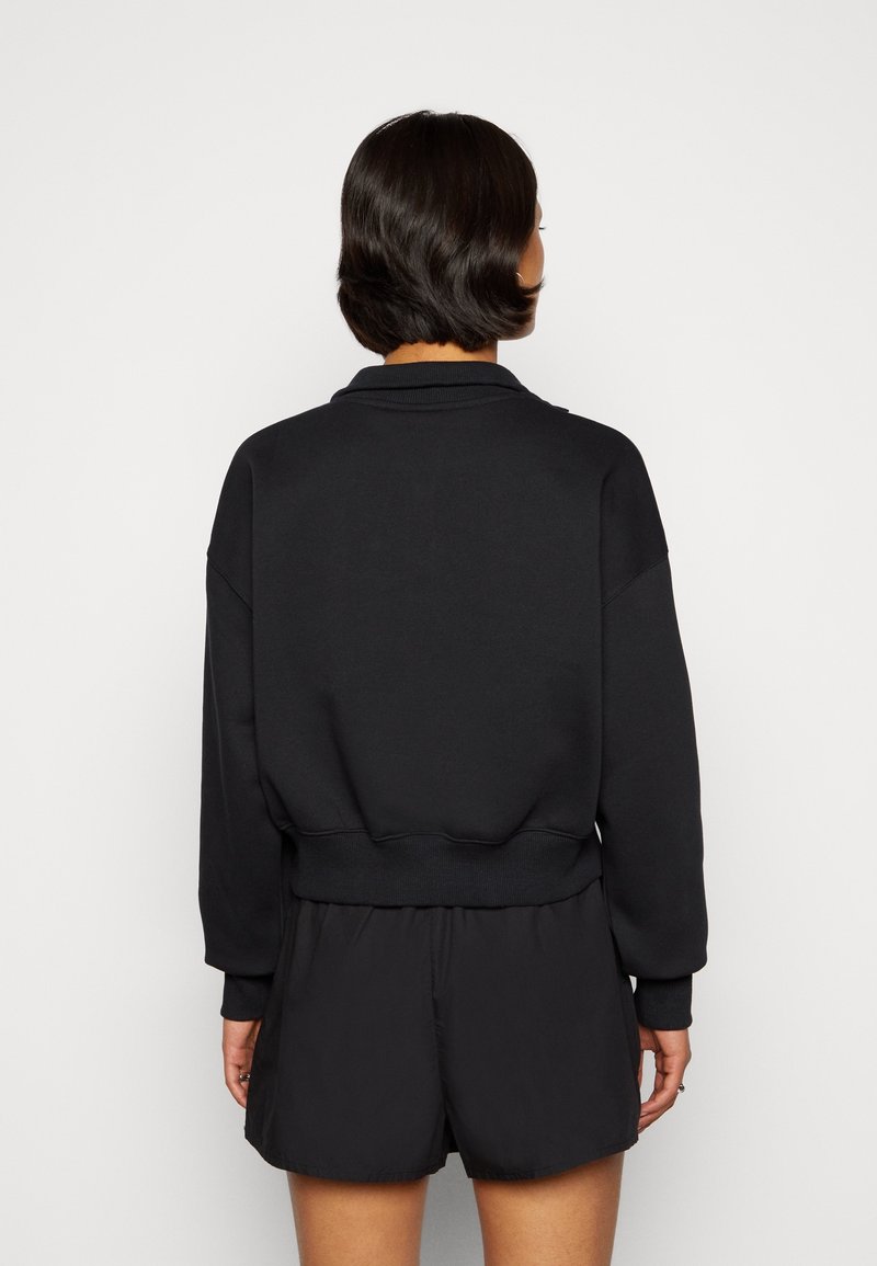 Vrouw met kort donker haar, gekleed in een zwart sweatshirt met lange mouwen en zwarte korte broek, gezien van achteren tegen een effen achtergrond.