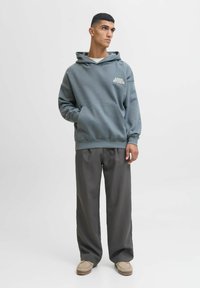 Mikina s kapucí v tlumené modro-zelené bavlně, s přední kapsou a textem "APPAREL STUDIOS". V kombinaci s volnými šedými kalhotami a béžovými botami.