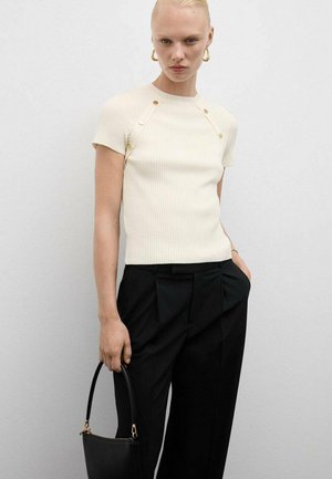 Crèmewit geribbeld gebreid top met korte mouwen en knoopaccenten, gecombineerd met hoge zwarte broeken en een kleine zwarte handtas.