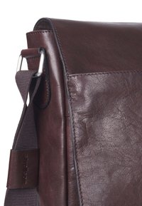 Bolsa mensajera de cuero marrón con herrajes plateados, superficie texturizada y correa de tela ajustable. Cuenta con un cierre de solapa y un bolsillo lateral.