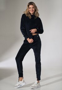 Marineblauwe velours trainingspak met lange mouwen en taps toelopende broek, met een ontspannen pasvorm en een subtiele glans. Gecombineerd met lichtgekleurde sneakers.