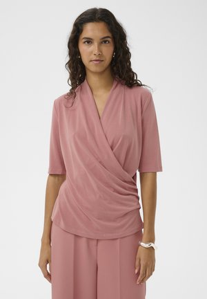Vrouw die een stofroze wikkelshirt en bijpassende brede broek draagt, met een zilveren armband om haar rechterpols, naar voren kijkend.