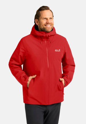 Lächelnder Mann, der eine rote Jack Wolfskin Kapuzenjacke trägt, mit Händen in den Taschen, vor einem schlichten hellen Hintergrund.